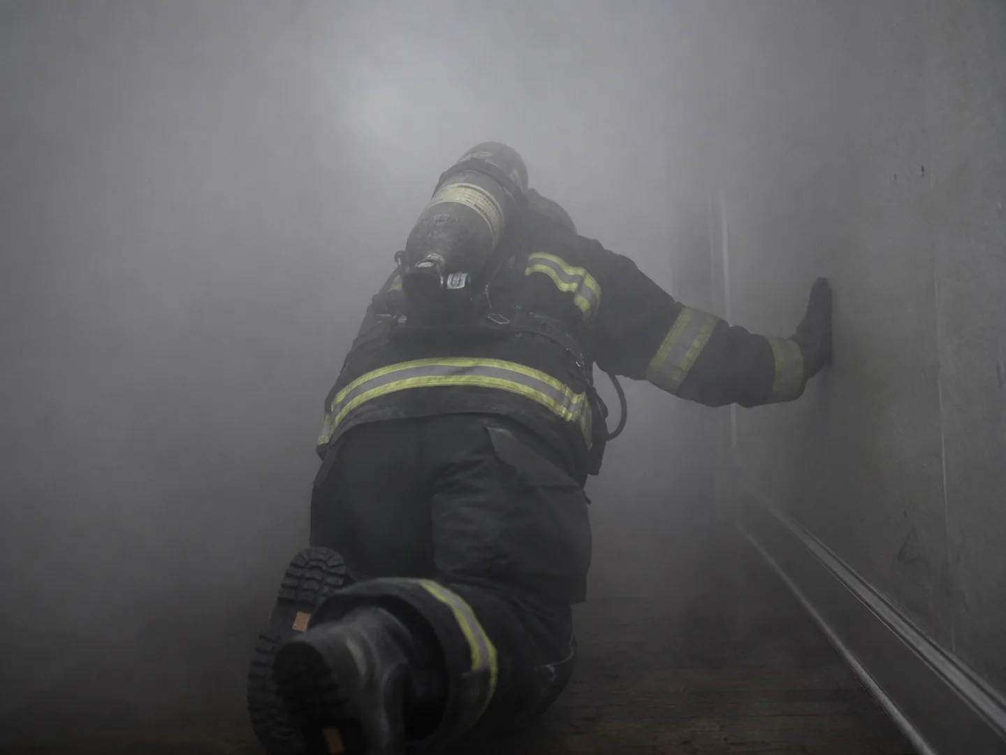 A firefighter in full turnout gear crawling through a smoke-filled hallway, one gloved hand feeling along the wall to navigate